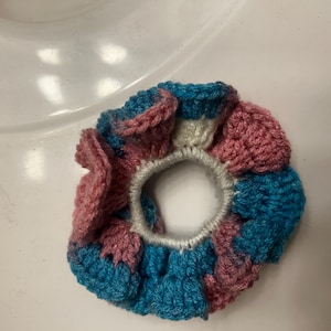 May include: A circular hair scrunchie featuring a crocheted design with alternating sections of dusty rose pink and vibrant blue yarn. The center of the accessory consists of a white elastic band, providing a gathered, ruffled texture throughout.