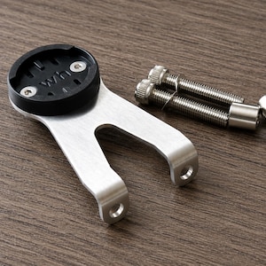 May include: A silver-colored bicycle computer mount with a black top section, accompanied by silver screws and a small cylindrical spacer. The mount has a curved design with two holes for attachment. The letters "WH" are visible on the black top.