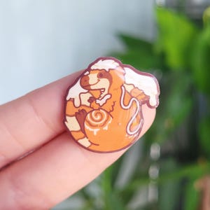 May include: A circular pin featuring an orange dinosaur character with a cream-colored swirl on its belly, resembling a cinnamon roll. The dinosaur has a white frosting-like topping on its head and back, with a glossy finish and dark brown outline.