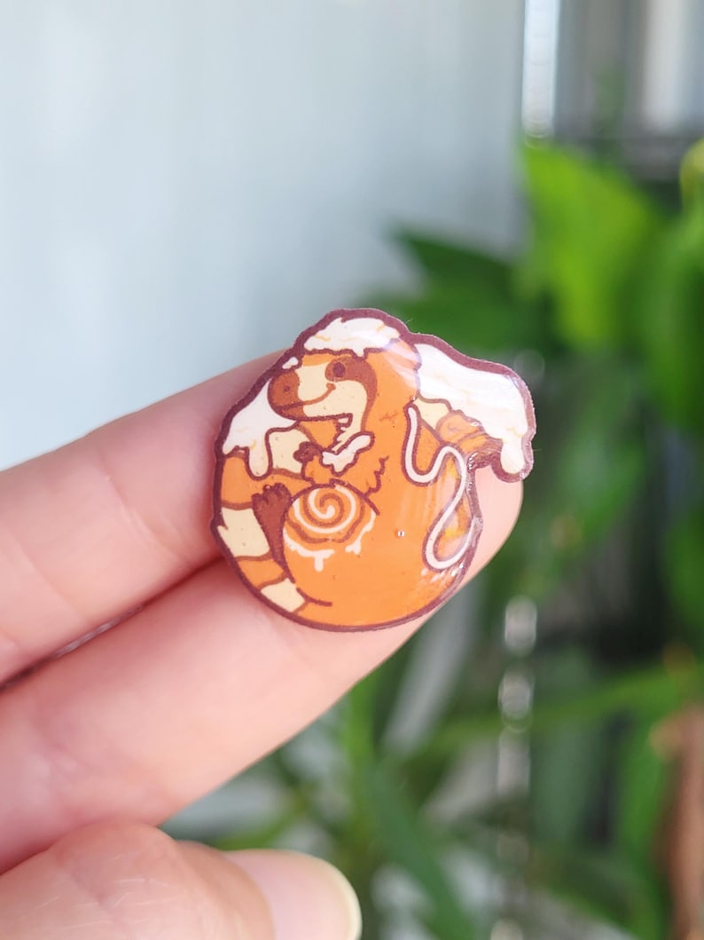 May include: A circular pin featuring an orange dinosaur character with a cream-colored swirl on its belly, resembling a cinnamon roll. The dinosaur has a white frosting-like topping on its head and back, with a glossy finish and dark brown outline.