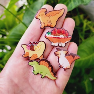 May include: A collection of six dinosaur-themed enamel pins. The pins feature various dinosaurs in cartoon styles, with colors including orange, green, and brown. Some pins have food-themed designs, such as a dinosaur with a strawberry cake.