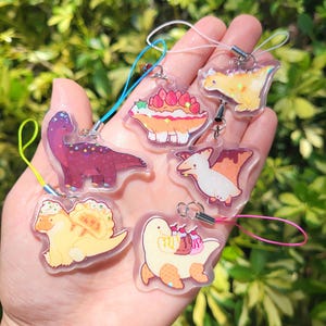 Puede incluir: Colección de siete colgantes de dinosaurios de acrílico transparente con diseños coloridos. Cada colgante presenta una ilustración de dinosaurio diferente con acentos temáticos de comida, como fresas y crema batida. Los colgantes tienen cordones adjuntos de varios colores.