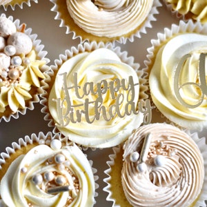 May include: A close-up of six cupcakes with white and beige frosting. The cupcakes are decorated with gold sprinkles, pearls, and a gold "Happy Birthday" topper.