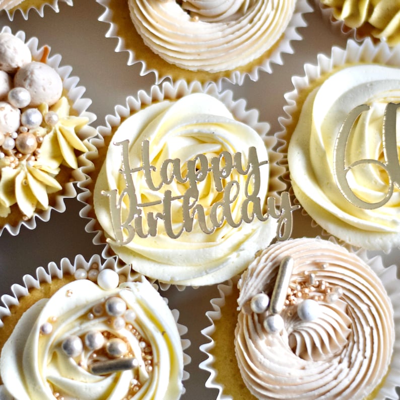 May include: A close-up of six cupcakes with white and beige frosting. The cupcakes are decorated with gold sprinkles, pearls, and a gold "Happy Birthday" topper.