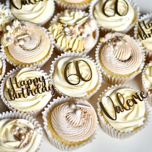 May include: A collection of cupcakes decorated with gold lettering and sprinkles. The cupcakes are frosted with white and beige frosting. Some of the cupcakes have the words "Happy Birthday" and "Valerie" on them. Other cupcakes have the number "60" on them.