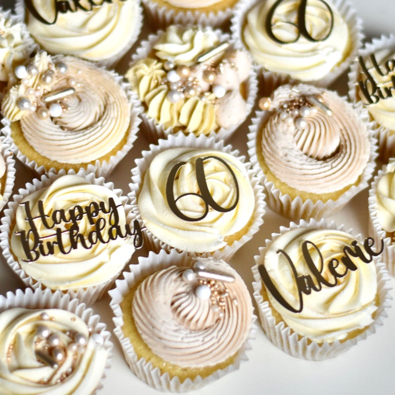 May include: A collection of cupcakes decorated with gold lettering and sprinkles. The cupcakes are frosted with white and beige frosting. Some of the cupcakes have the words "Happy Birthday" and "Valerie" on them. Other cupcakes have the number "60" on them.