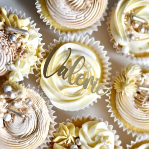 May include: A close-up of cupcakes decorated with white and gold frosting. One cupcake has a gold topper that says "Valerie".