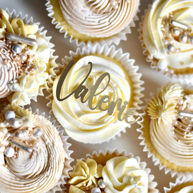 May include: A close-up of cupcakes decorated with white and gold frosting. One cupcake has a gold topper that says "Valerie".