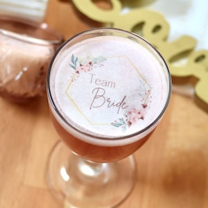 Puede incluir: Un vaso de bebida rosa con una pegatina blanca en la parte superior que dice "Team Bride" en letras doradas con un diseño floral alrededor.
