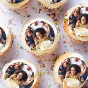 May include: A close-up of cupcakes decorated with edible images of a family celebrating a birthday. The cupcakes are white with a white frosting and are arranged on a white surface with colorful sprinkles.