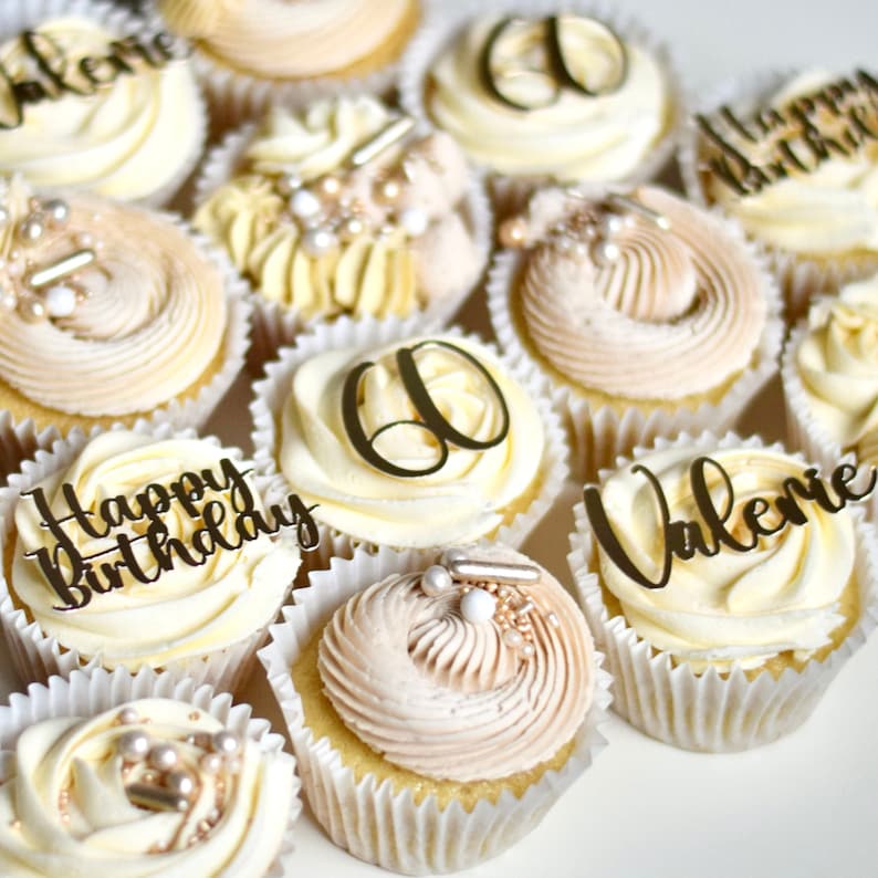 May include: A collection of cupcakes decorated with gold and white frosting. Some cupcakes have gold lettering that says "Happy Birthday" and "Valerie". Other cupcakes have a gold number "60" on top.