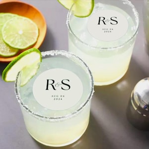 Può includere: Due cocktail margarita in bicchieri trasparenti con bordi salati, guarniti con spicchi di lime. Ogni bicchiere ha un adesivo circolare bianco con le lettere "R&S" e la data "04 AGO 2024". Sullo sfondo una ciotola di legno con fette di lime.