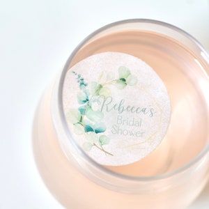 Peut inclure: Un autocollant rond blanc avec un cadre géométrique doré et des feuilles d'eucalyptus vertes en aquarelle. Le texte "Rebecca's Bridal Shower" est écrit en écriture cursive noire.