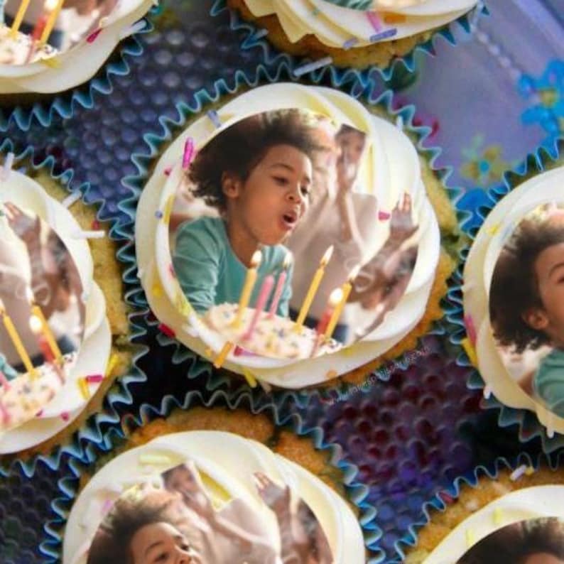 May include: A close-up of cupcakes decorated with edible images of a child blowing out candles on a birthday cake. The cupcakes are arranged in a circle on a blue and white patterned surface.