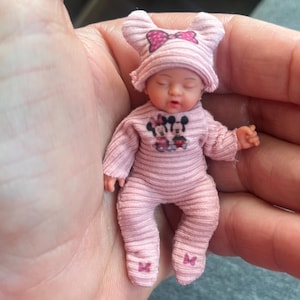 May include: A miniature baby doll wearing a light pink ribbed long-sleeve onesie and a matching hat with ear-like protrusions. The outfit features a small graphic of Minnie and Mickey Mouse on the chest, a pink bow on the hat, and small bow accents on the feet.