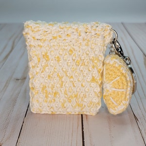 May include: A crocheted lemon-shaped keychain with a white and yellow color scheme. The keychain is attached to a small crocheted pouch with a zipper.