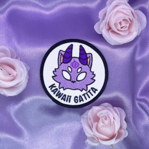 May include: Purple and white embroidered patch with a cartoon cat with a third eye and the text "Kawaii Gattita" around the edge.