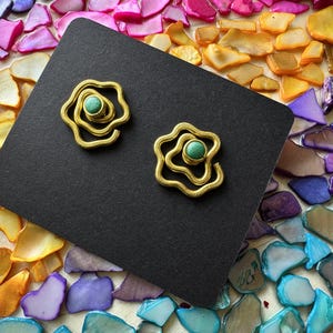 May include: A pair of gold-toned metal earrings featuring a swirling, organic wire design. Each earring is centred with a small, round, textured green stone. The earrings are displayed on a rectangular black card.