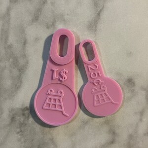 May include: Two pink plastic tokens. One token reads "$1" and the other reads "25¢". Both tokens have a circular design with a stylized image of a building. Each token has a loop at the top.