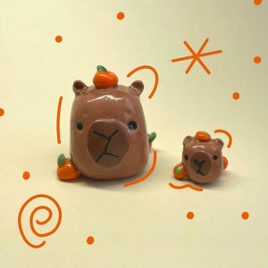 May include: Two brown ceramic figurines of capybaras wearing orange pumpkin hats. The larger capybara is sitting with its legs outstretched, while the smaller capybara is standing. Both capybaras have black eyes and noses.