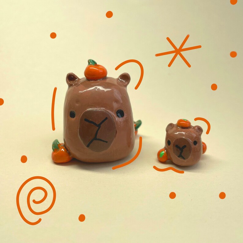 May include: Two brown ceramic figurines of capybaras wearing orange pumpkin hats. The larger capybara is sitting with its legs outstretched, while the smaller capybara is standing. Both capybaras have black eyes and noses.