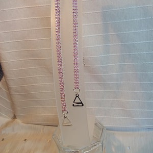 May include: Two pink rhinestone bra straps with silver metal clasps. The straps are hanging on a white stand.