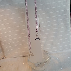 May include: Two sets of bra straps with pink rhinestones. The straps are displayed on a clear, rectangular stand. Each strap has a silver-coloured metal clasp. The background is a white, textured fabric.
