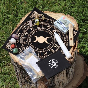 Peut inclure: Un assortiment d'articles spirituels disposés sur une souche d'arbre. L'ensemble comprend un tissu d'autel noir avec un motif de zodiaque, des cristaux, une bougie blanche, une petite boîte avec un pentagramme et un paquet de sauge blanche.
