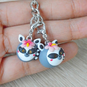 May include: A silver keychain with two gray and white lemur charms. Each lemur charm has a pink flower with a yellow center and black eyes.