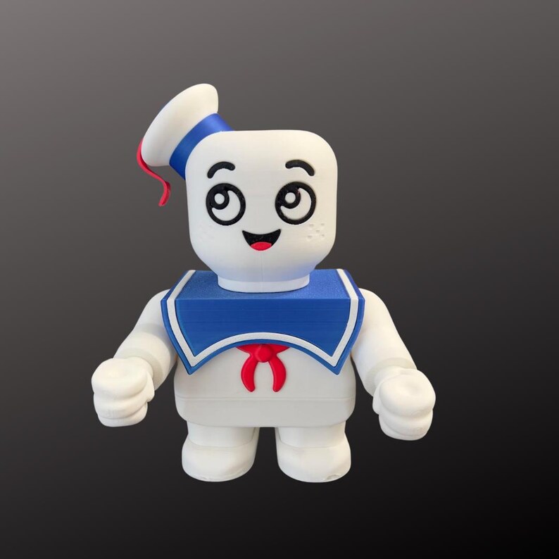 May include: A white figurine of the Stay Puft Marshmallow Man, featuring a sailor hat with blue and red accents, a blue collar, and a red bow tie. The figure has a smiling face with black eyes and a red mouth.
