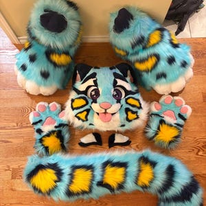May include: A complete fursuit set in a blue, yellow, and black color scheme. The set includes a head, paws, feet, tail, and eyelids. The head features a cartoonish design with large eyes and a protruding tongue. The paws and feet have a paw pad design.