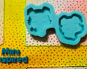 May include: Two blue silicone molds for making resin crafts. Each mold features a cartoon animal with a smiling face. The molds are on a yellow background with a ruler.