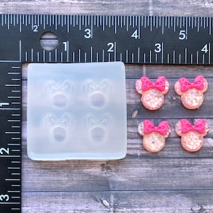 May include: A clear silicone mold with four cavities shaped like Minnie Mouse heads. The mold is next to four finished resin Minnie Mouse head charms with pink bows and glitter.