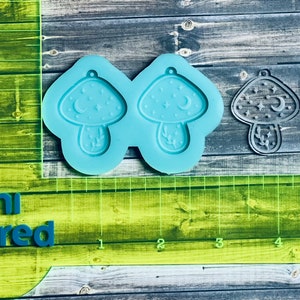 May include: A blue silicone mold with two mushroom shapes. Each mushroom has a crescent moon, stars, and leaves. The mold is on a wooden surface with a green ruler.