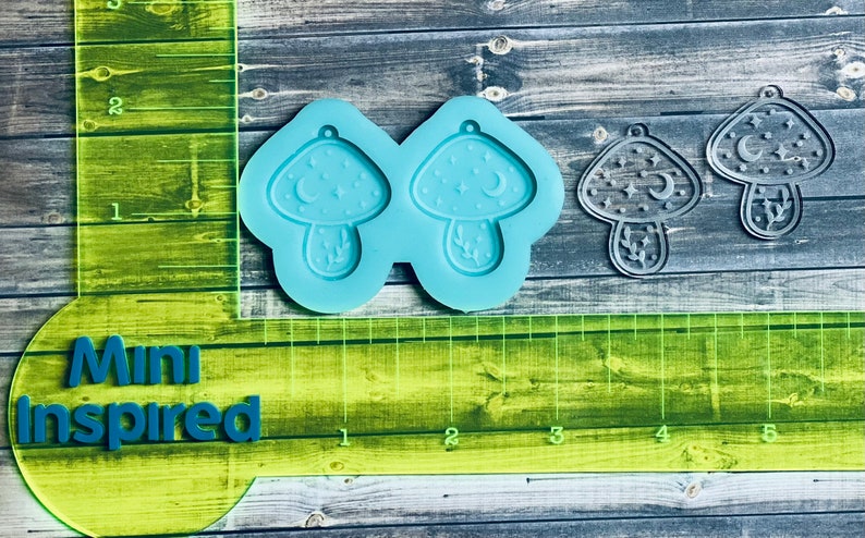 May include: A blue silicone mold with two mushroom shapes. Each mushroom has a crescent moon, stars, and leaves. The mold is on a wooden surface with a green ruler.