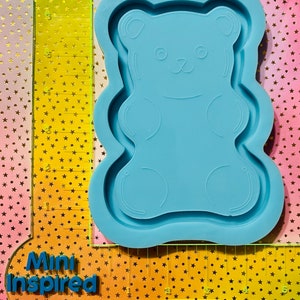 May include: A light blue silicone mold shaped like a gummy bear. The mold is approximately 5 inches tall and 4 inches wide. The mold has a detailed design of a gummy bear with a smiling face and small arms and legs.