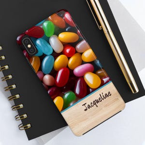 May include: A phone case with a colorful jelly bean design and a wood-grain background. The name "Jacqueline" is written on the wood-grain background.