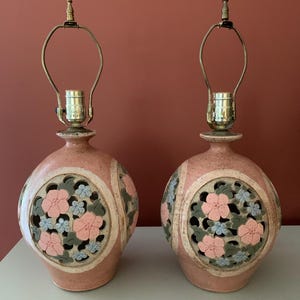 May include: A pair of spherical ceramic table lamps with a speckled pink finish. Each lamp features a circular cutout panel displaying a relief of pink flowers, blue blossoms, and green leaves. The lamps include brass-toned hardware and sockets.