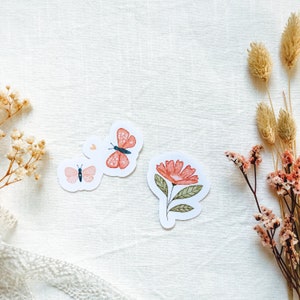 Könnte beinhalten: Drei Aufkleber mit einem rosa Schmetterling, einem roten Schmetterling und einer rosa Blume mit grünen Blättern. Die Aufkleber befinden sich auf einem weißen Hintergrund.