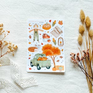 Può includere: Un foglio di adesivi a tema autunnale con illustrazioni di uno spaventapasseri, zucche, un camioncino pieno di zucche, una torta, una lanterna e un albero. Gli adesivi sono su uno sfondo bianco con uno striscione rosa che dice "SOMEKINDART by Julie Lee".