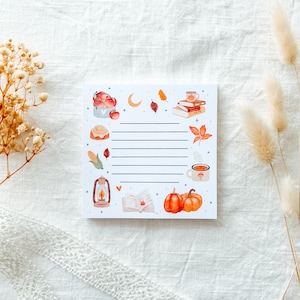 Könnte beinhalten: Ein quadratischer, linierter Notizblock mit herbstlichen Illustrationen. Das Design umfasst Kürbisse, Blätter, Bücher, eine Laterne und eine Tasse Tee. Der Notizblock ist weiß mit einem dekorativen Rand aus Herbstmotiven.