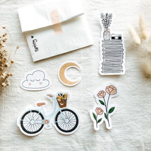 Könnte beinhalten: Ein Set mit Aufklebern, die ein Fahrrad mit Blumenkorb, eine Wolke mit einem Smiley, einen Halbmond, einen Stapel Bücher mit einer Topfpflanze und einer Kamera darauf sowie zwei rosa Blumen zeigen.
