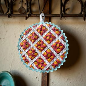 May include: Round quilted hot pad with a white lattice design over a red and yellow floral pattern. The edge is trimmed with a light blue scalloped border and a white hanging loop. The hot pad is hanging on a metal rack.