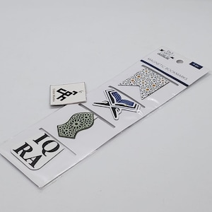 Puede incluir: Un conjunto de cinco marcapáginas magnéticos con patrones geométricos islámicos y caligrafía. Los marcapáginas son blancos con detalles azules, verdes y dorados. El texto en los marcapáginas dice "IQRA".