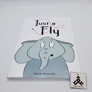 May include: A white book with a blue elephant illustration on the cover. The title of the book is "Just a Fly" and the author is "Saajida Rhemtullla".