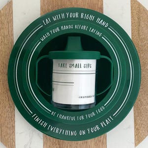 May include: A green plastic plate with white text that reads "Eat with your right hand, wash your hands before eating, take small sips, be thankful for your food, finish everything on your plate". A clear plastic cup with a green lid and handle is in the center of the plate.