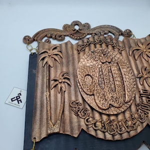 May include: A decorative wall hanging with a copper-colored finish. The hanging features an intricate design of palm trees, Arabic calligraphy, and a large, circular "Allah" inscription. The hanging is attached to a black backing with a scalloped edge.