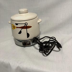 May include: Off-white ceramic fondue pot with a lid and side handles. The pot features a retro design with a yellow fork, brown fish, and black utensils. It sits on a chrome base with a black power cord.