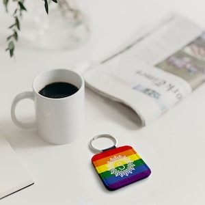Może przedstawiać: Brelok skórzany w kolorze tęczy z białym wzorem mandali i literą "S" w środku. Brelok leży na białym tle obok filiżanki kawy i gazety.