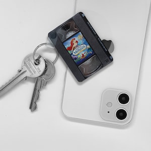 May include: A white smartphone with a black VHS tape case attached to it with a silver key chain. The VHS tape case has a picture of Ariel from The Little Mermaid on it. The text on the VHS tape case reads "The Little Mermaid".